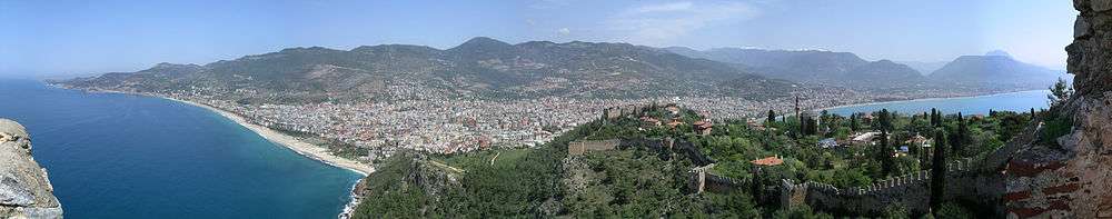 A panoramic view of a city beneath a mountain range with blue sea on both sides of a peninsula. On the peninsula is a castle wall and red roofed buildings. A young girl peers into the scene over the wall on the far left.