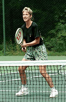 A blonde-haired female tenis player with multi-colored shorts and a black shirt, with the tenis racket out in front of her