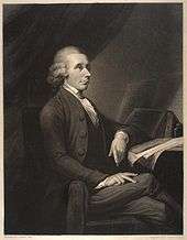 Portrait of a man sitting on a chair and leaning against a table with books and papers.