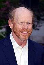 Portrait of a balding, red-haired Caucasian male who is wearing an unbuttoned white collared shirt over a blue suit.