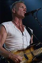 Adam, beyaz kesim bir gömlek giymiş ve gitar çalarken şarkı söylüyor..