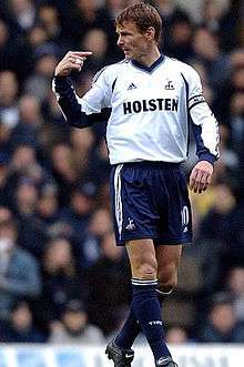 Teddy Sheringham - wearing a long-sleeved white jersey, dark blue shorts and a Premier League captain's armband on the left arm - points at himself.